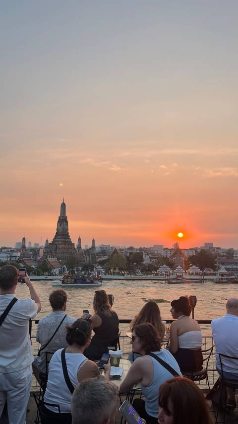Fiery sunset descends behind the spires of Wat Arun and Bangkok skyline while the sun glows orange above the Chao Phraya River.
