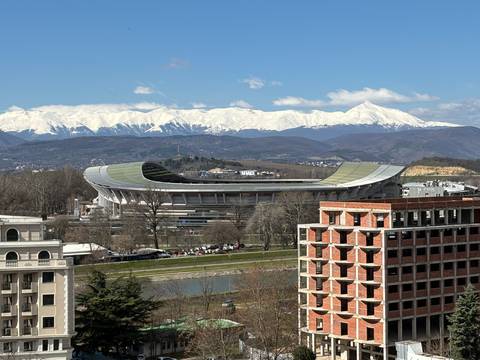 Modern oval stadium set against a river and distant snow-capped mountain range on a clear day.