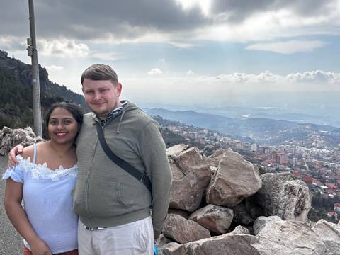 Young couple posing on a rocky mountain viewpoint overlooking a sprawling city and distant ridges under a partly cloudy sky.