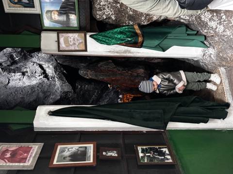 Small child in a striped beanie stands at the entrance of a dim rock grotto adorned with framed photos and green velvet drapes.
