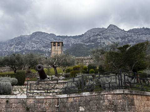 Historic cannon set among manicured shrubs with a stone tower and rugged mountains rising behind under overcast skies.