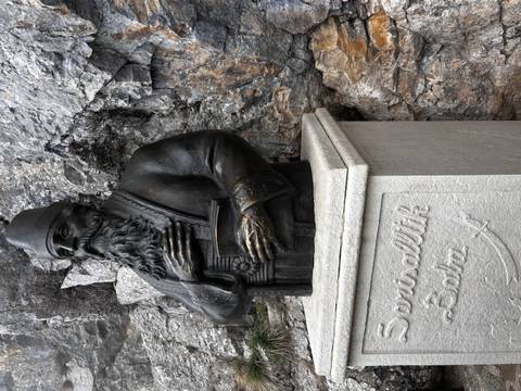 Bronze bust of a robed man holding a book, mounted on a carved stone pedestal against a rocky cliff.