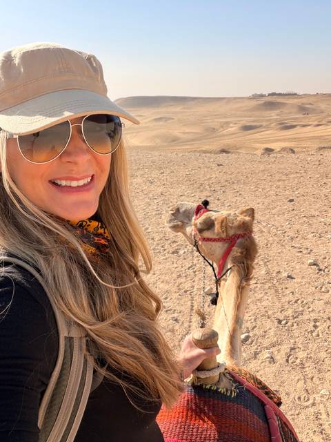 Woman taking a selfie with a saddled camel in a vast desert landscape