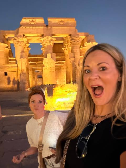 Two women posing playfully in front of a dramatically lit ancient Egyptian temple at night