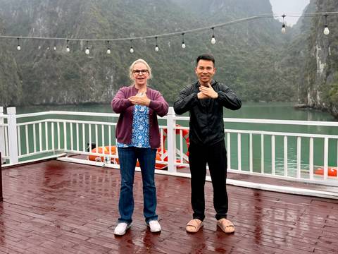 Tourist and guide practise Tai-chi gestures on a wet cruise deck with limestone peaks behind.