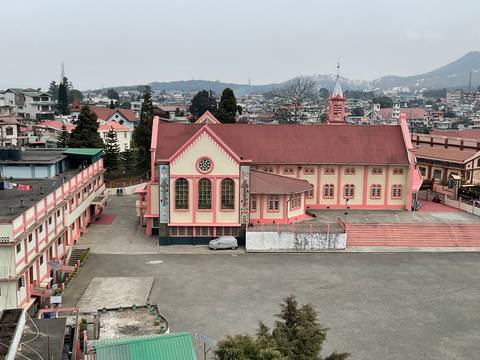 A pink-roofed church with tall arched windows overlooks a hill town dotted with buildings and distant mountains.