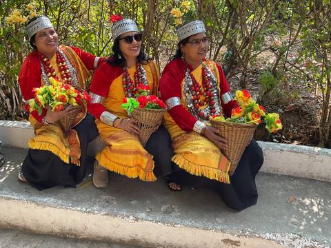 Three women in vibrant traditional Meghalayan attire sit holding baskets of colorful flowers.