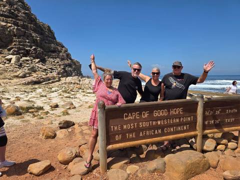 Travelers posing at the Cape of Good Hope sign beside rocky coast and waves.