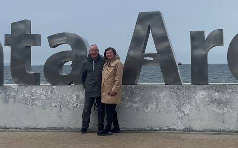 Couple standing in front of large metal Punta Arenas seaside letters with grey ocean backdrop.