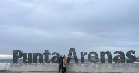 A couple poses in front of the large metallic Punta Arenas sign beside the sea under an overcast sky.