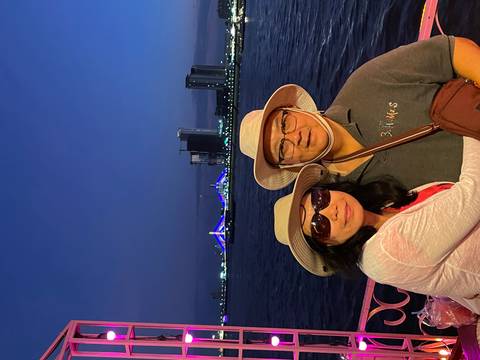 Smiling couple wearing sunhats on a night river cruise with a brightly lit suspension bridge in the background.