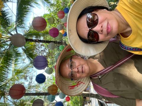 Selfie of two travelers beneath colorful hanging lanterns and palm fronds in a tropical setting.