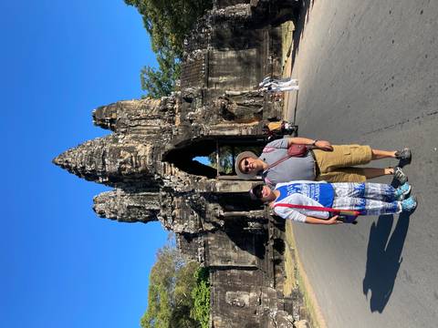 Couple posing on a roadway before the towering stone South Gate of Angkor Thom under a clear blue sky.