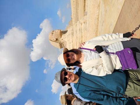 Smiling couple poses in front of the Great Sphinx of Giza under a bright blue sky and fluffy clouds.