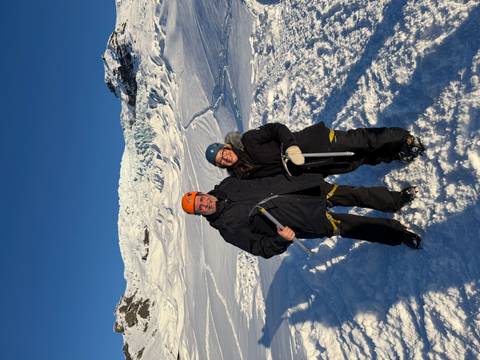 Two travellers in snow gear and helmets stand on a glacier with ice axes, bright sun illuminating the icy slope behind.
