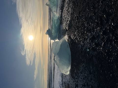 Translucent icebergs glitter on a black-pebble beach at sunrise with the sun low over the Atlantic waves.