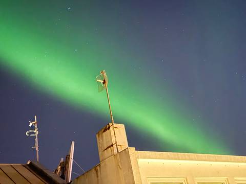 Vivid green aurora ribbon across a starry night sky above a rooftop with antennas.