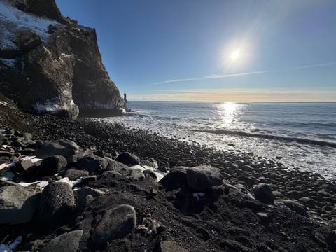 Sunlit winter seascape with black-pebble beach, basalt sea stack and shimmering ocean horizon.