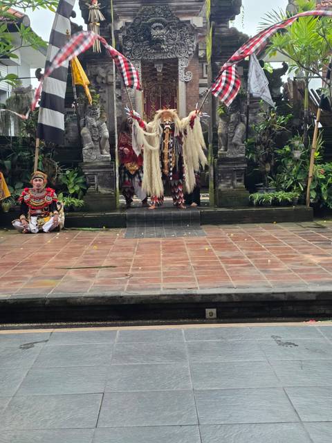 Balinese performers in elaborate costumes act on a temple stage flanked by stone carvings and greenery.