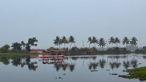 Traditional Kerala houseboat glides on calm backwaters lined with palm trees under a hazy sky.