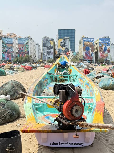 Colorful wooden fishing boat rests on a sandy urban beach with mural-covered buildings behind.