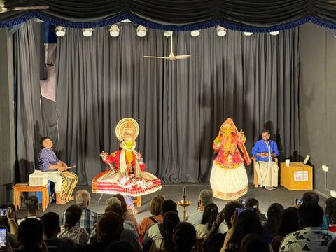 Kathakali dancers in elaborate costumes perform on a small theatre stage before an audience.