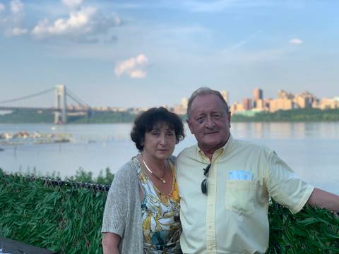 Older couple poses by a calm river with city skyline and suspension bridge behind.