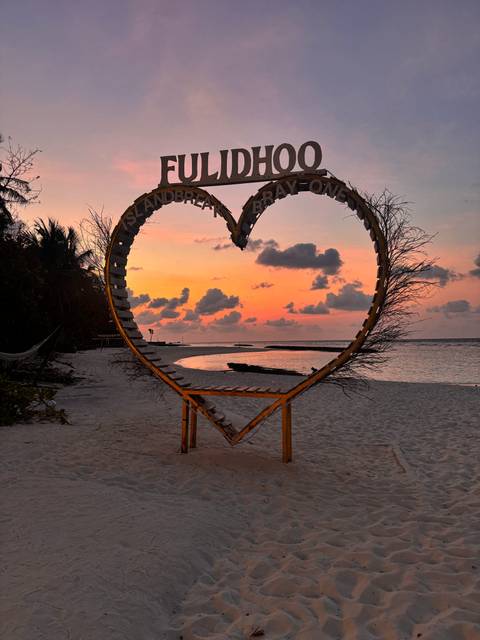 Heart-shaped wooden frame on a tropical beach silhouettes against a vivid orange sunset sky.