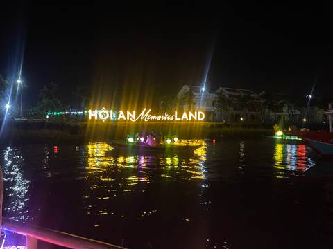 Glowing HOI AN Memories Land sign reflects in the river as tourists glide past in a lantern-lit boat at night.