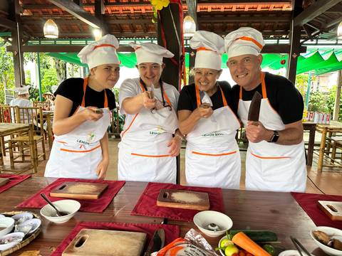 Cooking-class participants in white aprons and chef hats brandish knives over wooden boards, smiling broadly.