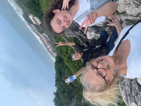 Three friends take a cheerful selfie on a rocky lookout high above a coastline and forested hills.