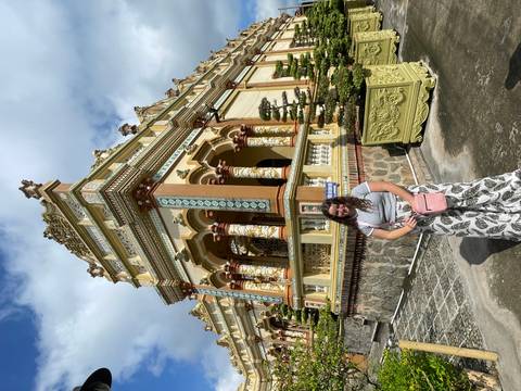 A traveller poses in front of an ornate, colourful pagoda decorated with detailed columns under bright sunlight.