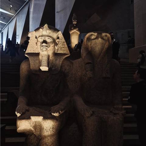 Dimly lit museum hall displaying twin ancient statues with visitors in the background