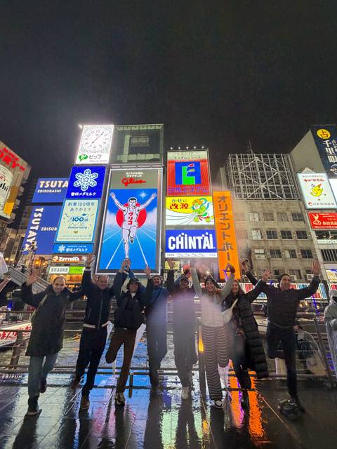 Smiling tour group posing with raised hands in front of Osaka's brightly lit Dotonbori billboards including the famous Glico running man.
