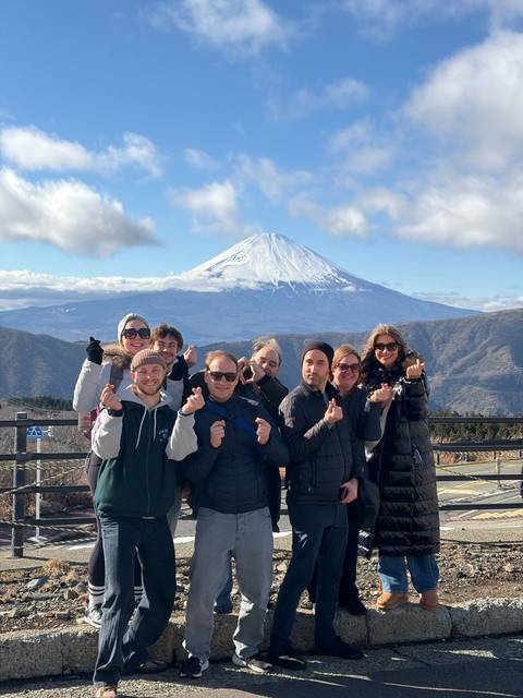 Tour group gesturing heart signs with snow-capped Mount Fuji towering in the sunny background.