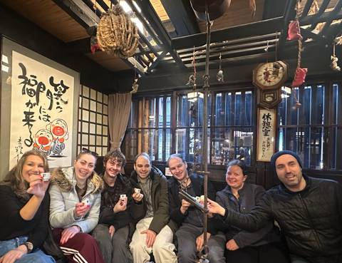 Friends seated inside a traditional Japanese house holding up lucky charms under wooden beams and hanging decorations.