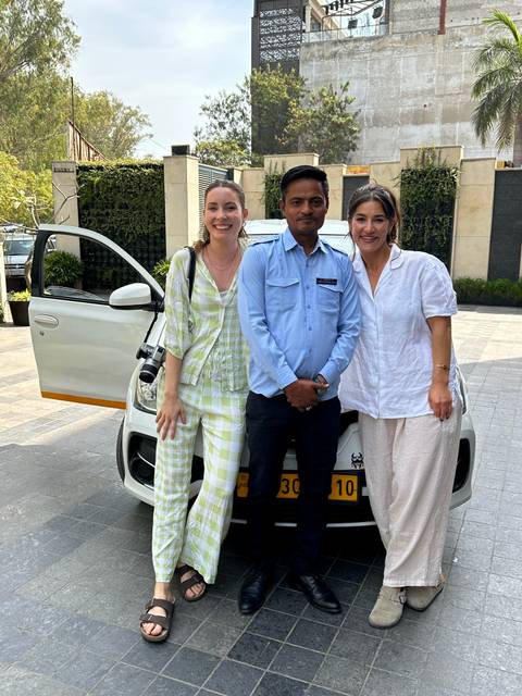 Two travellers pose with their driver in front of a white taxi outside a hotel entrance.