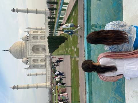 Visitors admire the Taj Mahal from its reflecting pool while two women sit foreground with backs turned.
