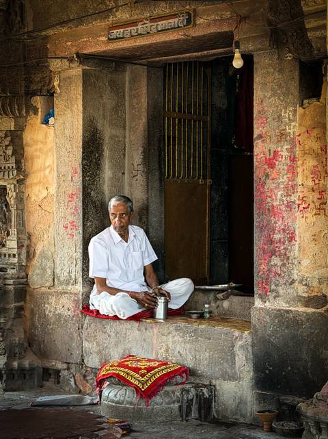 Elderly man in white clothing sits peacefully at the carved entrance of an Indian temple.