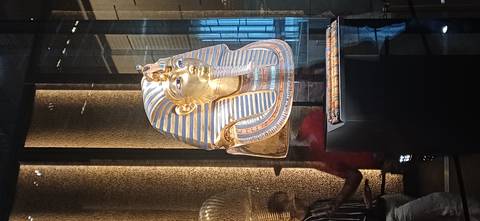 Gold funerary mask of Tutankhamun displayed behind glass inside a dimly lit museum gallery.