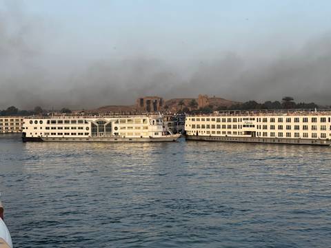 Two large Nile cruise ships docked side-by-side on a hazy morning with Luxor temple ruins in the distance.