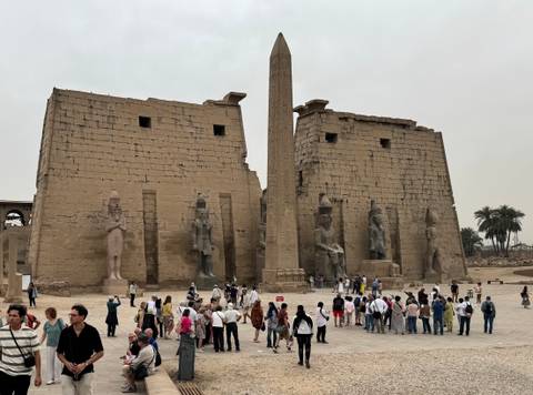 Crowds gather before the massive pylons and central obelisk of Luxor Temple under a grey sky.