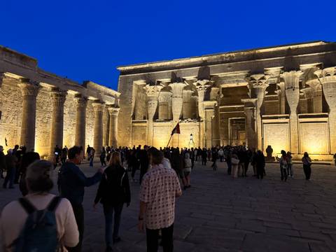 Evening crowd exploring the columned hall of Kom Ombo Temple illuminated in warm lights against a deep blue sky.