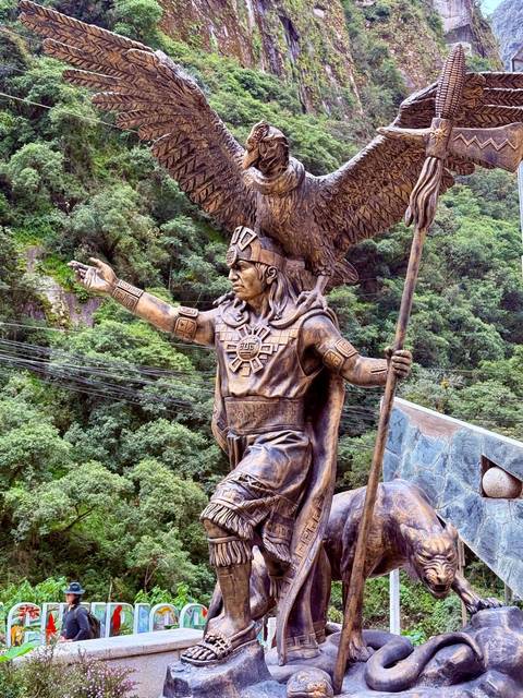 Bronze statue of an Inca warrior with a condor on his shoulder set against lush green forest in Aguas Calientes.