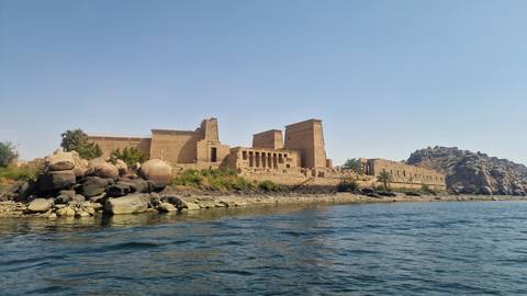 Ancient Temple of Philae rises above rocky Nile island with calm blue water in foreground.