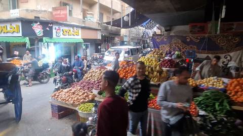 Busy Egyptian street market lined with colorful produce stalls and locals on motorbikes in motion blur.