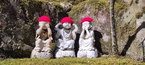 Three small stone monk statues wearing bright red knitted caps enact the ‘see no evil, hear no evil, speak no evil’ poses amid moss-covered rocks.