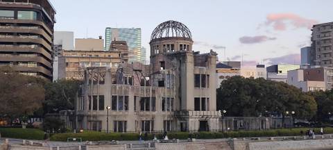 Twilight view of the ruined Atomic Bomb Dome on the riverbank with city buildings behind it.