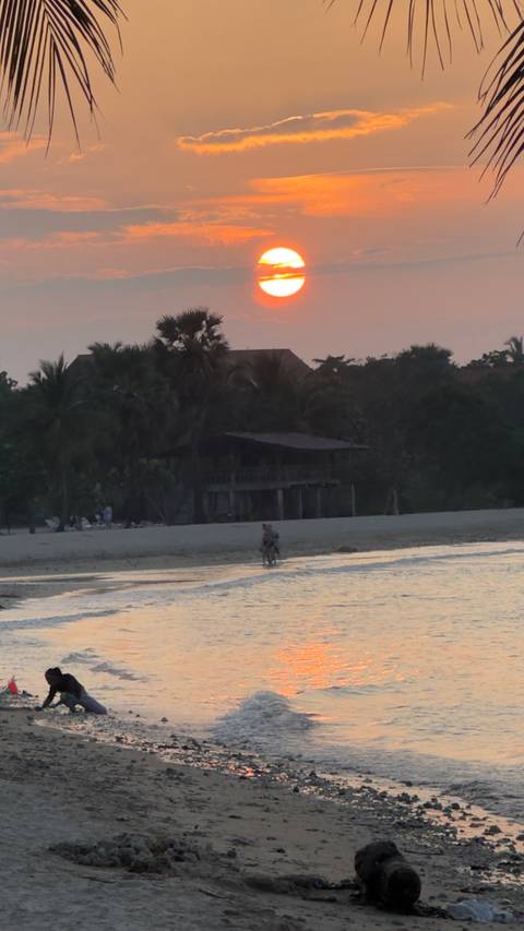 Two people cycling along a quiet beach at dusk with palm trees and a rustic wooden house behind.