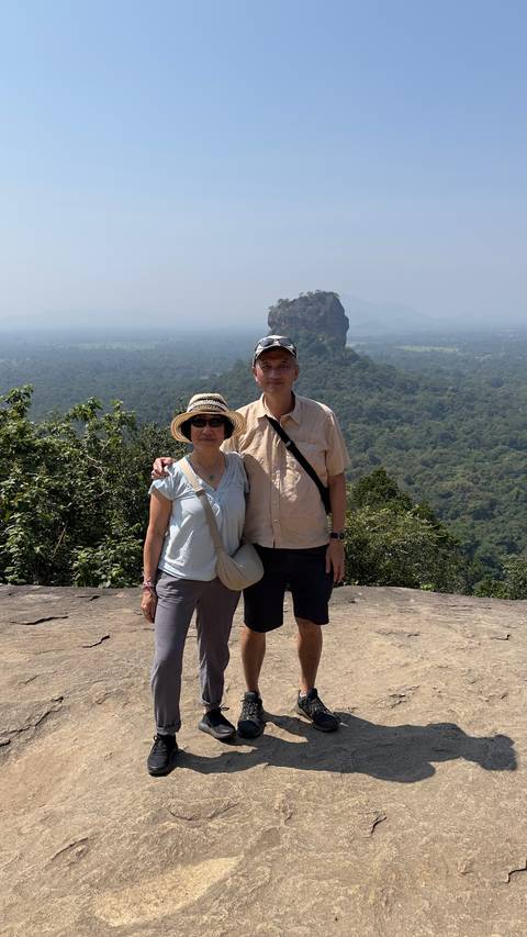 A couple stands arm-in-arm on a rocky outcrop overlooking lush Sri Lankan forest and a distant rock fortress.